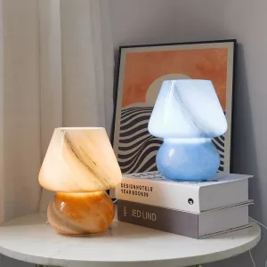 Modern glass mushroom table lamp with a warm light on, sitting on a wooden nightstand with white bedding in the background.