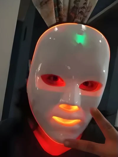 White LED facial beauty mask worn on a face showing red illuminated eye openings, an orange illuminated nose and mouth section, a green indicator light on the forehead, and an orange light ring glowing along the outer rim.