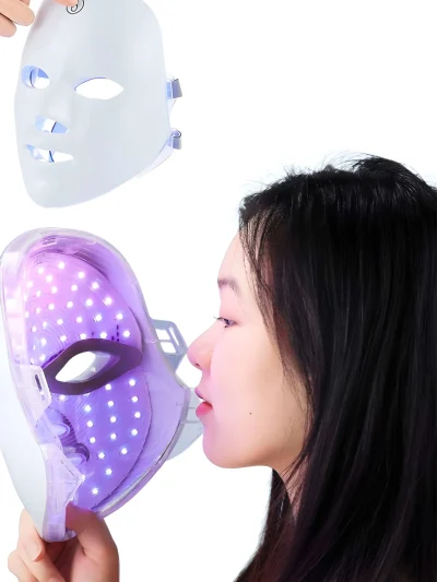 White LED facial beauty mask with purple light array held near a woman's face alongside a second white face mask showing a power button at the top.