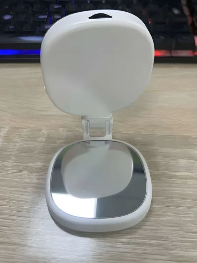White folding magnetic selfie light with mirror base resting open on a wooden table, featuring a square white light panel attached by a hinged joint to a mirror plate.