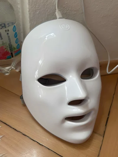 White glossy LED facial beauty mask with eye, nose, and mouth cutouts, featuring a power button icon on the forehead and a USB charging cable attached at the top.