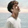 A young man with curly brown hair and closed eyes, wearing a white sweater and a white wireless earbud, stands against a backdrop of a calm ocean and a light sky.